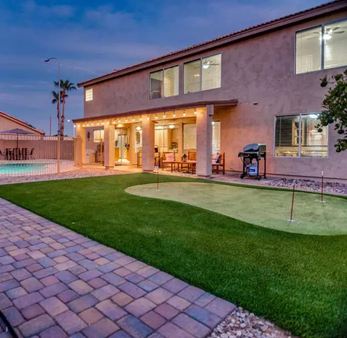 A backyard in Chandler, AZ features a quality putting green installed by expert Artificial Putting Green Installers, a grill, and a seating area under a covered patio adjacent to a two-story house at dusk. The yard is bordered by a stone walkway and fence, with a swimming pool on the left side.