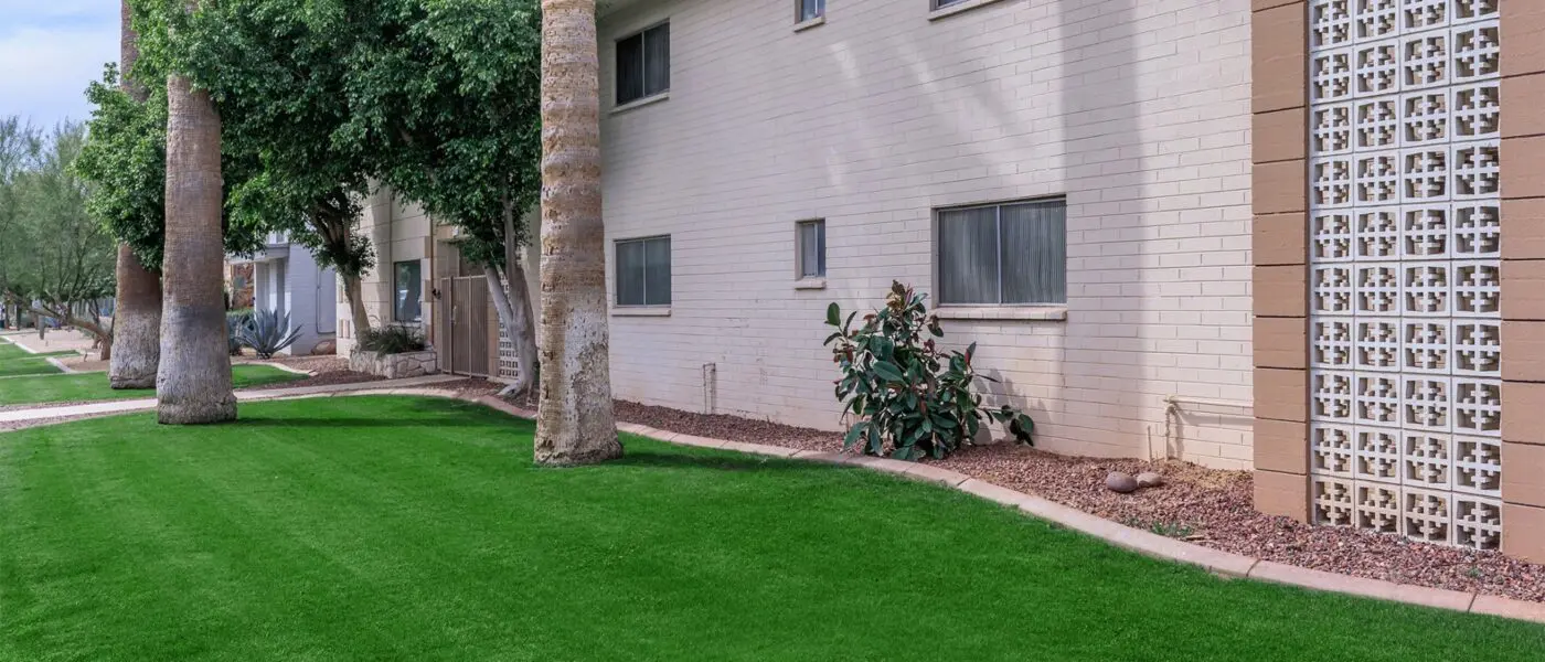 A well-maintained grassy lawn, complete with home putting greens, is adjacent to a beige brick building. The area features several rectangular windows and decorative block panels. Tall palm trees stand in contrast while shrubs and small plants line the base of the building for added charm.