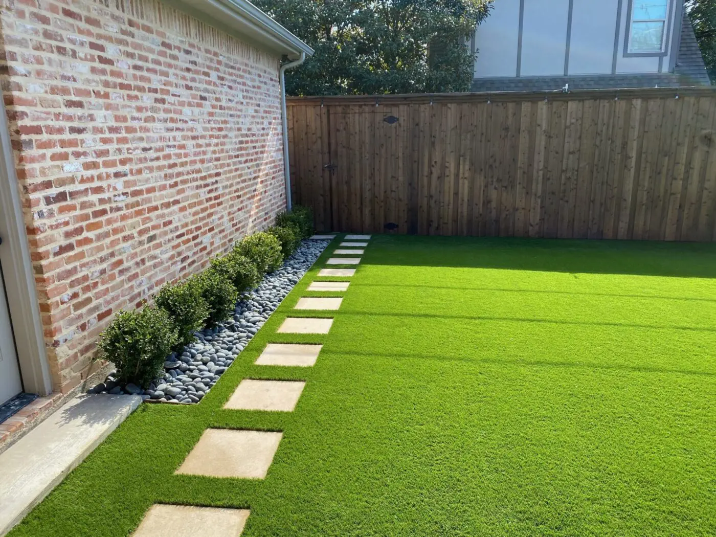 A well-maintained backyard featuring a brick wall on the left, a wooden fence in the background, and lush green pet turf. A row of small shrubs with a bed of white and gray pebbles runs alongside the wall. Square stepping stones create a path through the grass.