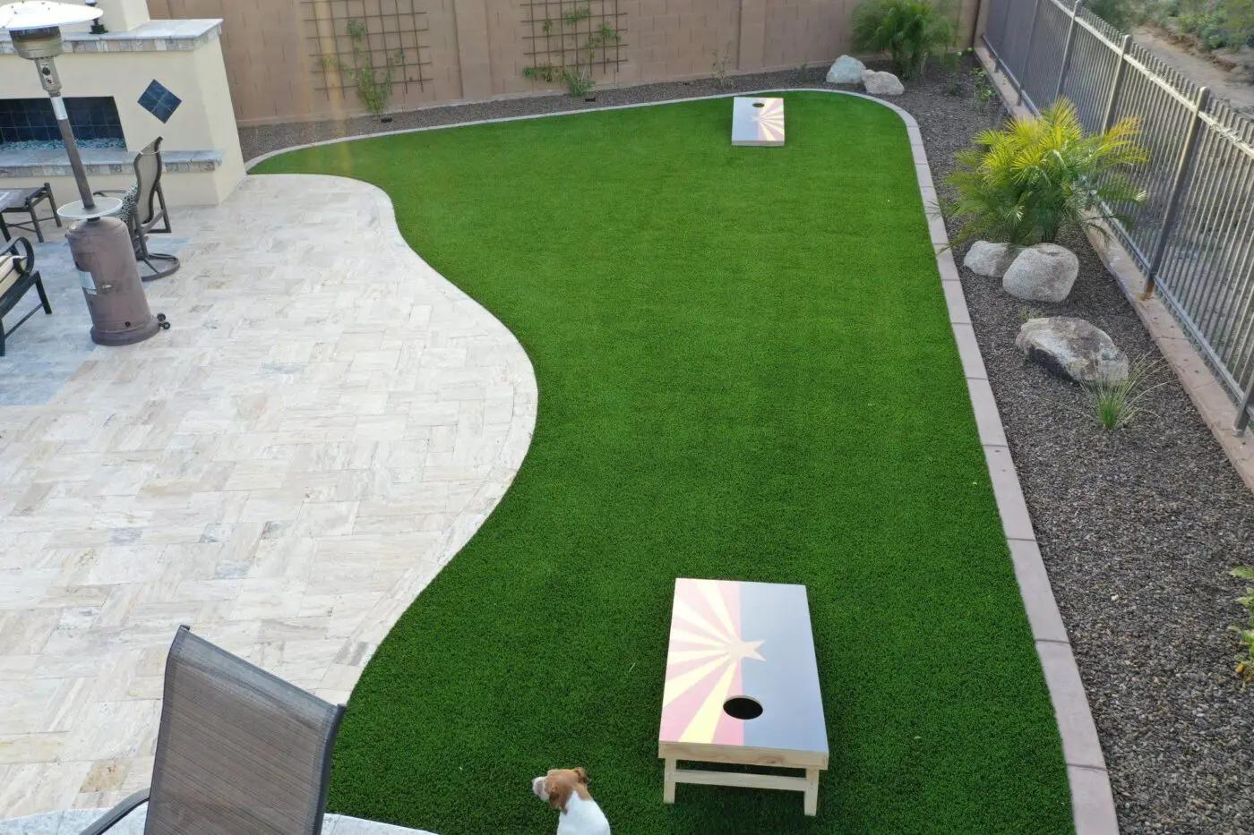 A backyard by Landscape Pros features a neatly manicured artificial grass lawn with two cornhole boards set up. There's a tiled patio with a chair and a dog in the lower left corner. The East Valley yard is bordered by a stone wall and metal fence, with decorative plants and rocks completing the serene setting.