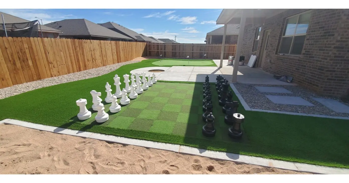 A backyard featuring a large outdoor chessboard with life-sized black and white pieces on pet turf. Adjacent is a paved patio area, surrounded by a wooden fence and various landscaping elements, including gravel and additional sections of waterless lawns.