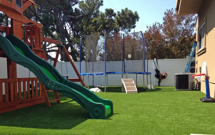A backyard setup features a green slide attached to a wooden play structure on the left, a trampoline with a safety net in the center, a swing near the middle, and a basketball hoop on the right. The yard, installed by Queen Creek's Artificial Grass Experts, showcases waterless lawns and is enclosed by a white fence.