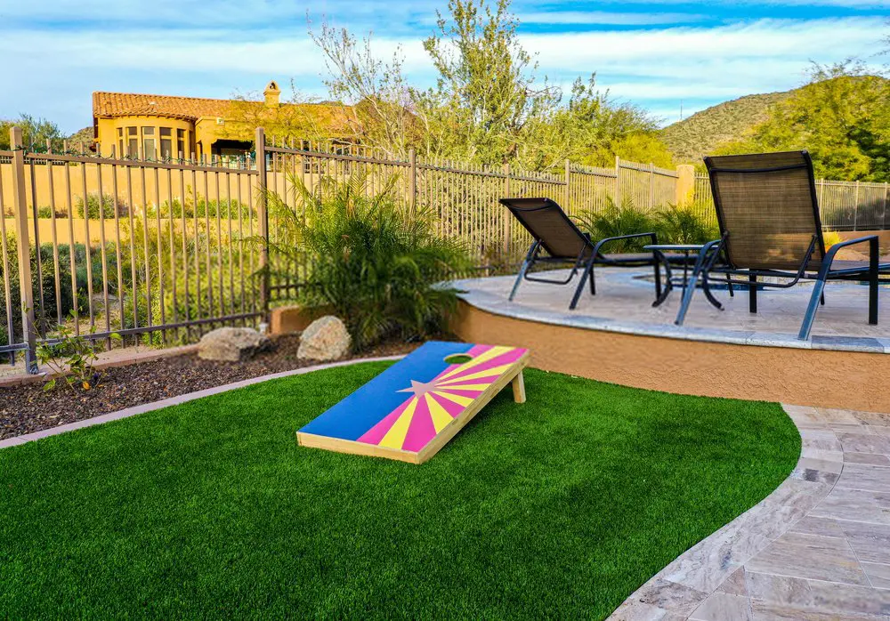 A backyard scene with a green artificial lawn installed by a professional artificial grass installer, a cornhole board featuring a vibrant design, and a patio area with black reclining chairs. A fenced enclosure surrounds the area with a scenic view of hills and a house in the background under a lightly clouded sky.