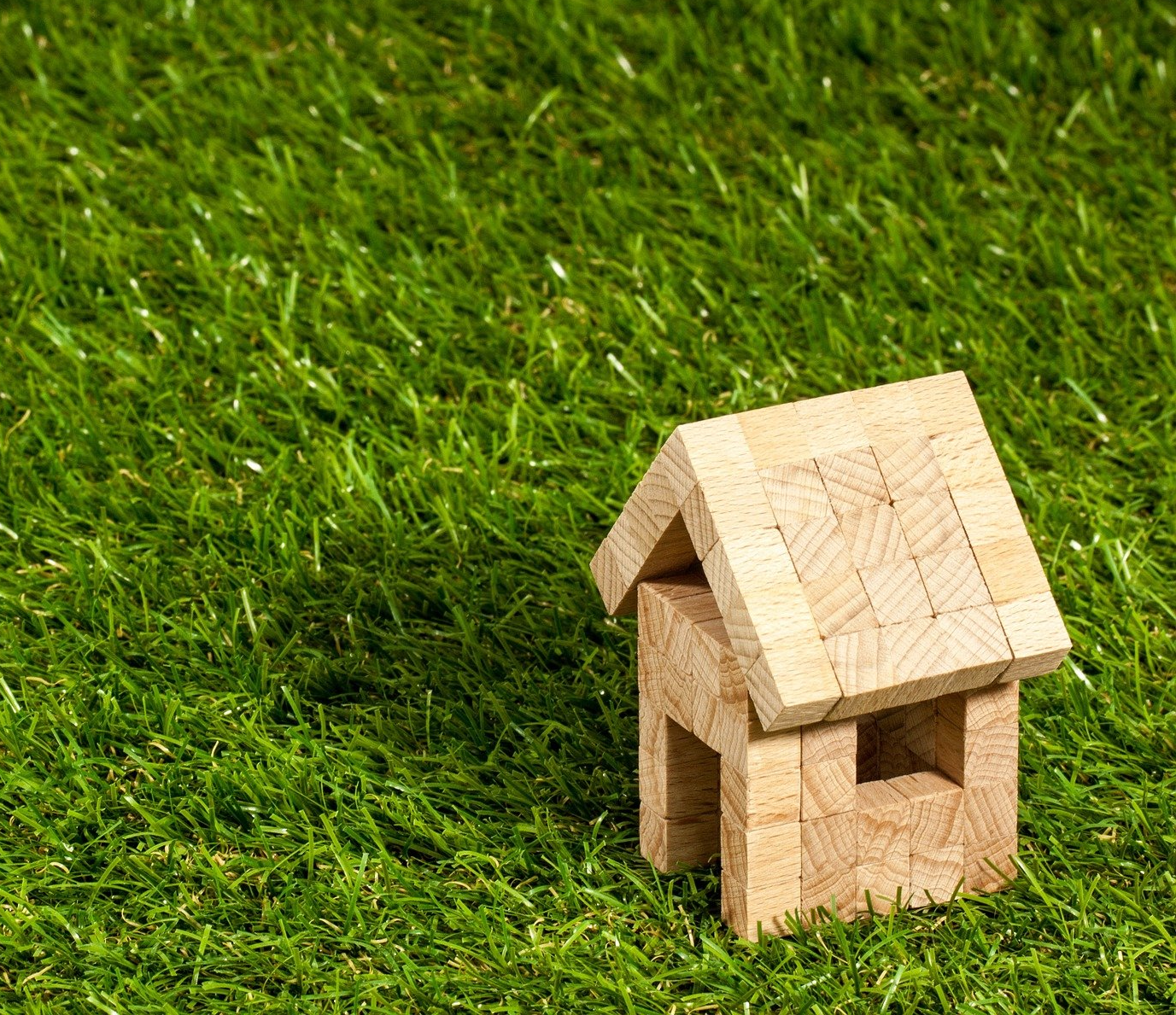 A small, simple model of a wooden house with a gabled roof sits on vibrant artificial grass, reminiscent of the installations in Tempe AZ. The house is made from square wooden blocks, giving it a minimalist and handcrafted appearance. The lush grass is evenly spread in the background.
