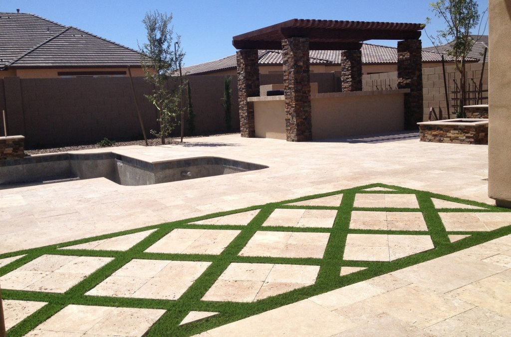 A backyard patio features beige stone tiles with green grass lines forming a diamond pattern. In the background, an unfinished pool sits beside a pergola-covered outdoor kitchen area with a stone bar. The scene is bordered by a tall privacy fence and enhanced with pet turf for easy maintenance.