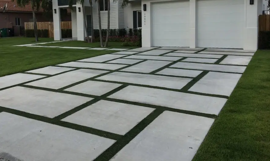 A modern driveway featuring large rectangular concrete slabs interspersed with artificial grass strips, leading up to a double garage with white doors. The surrounding lawn is well-manicured, and the house exterior is contemporary in design with white walls and large windows.