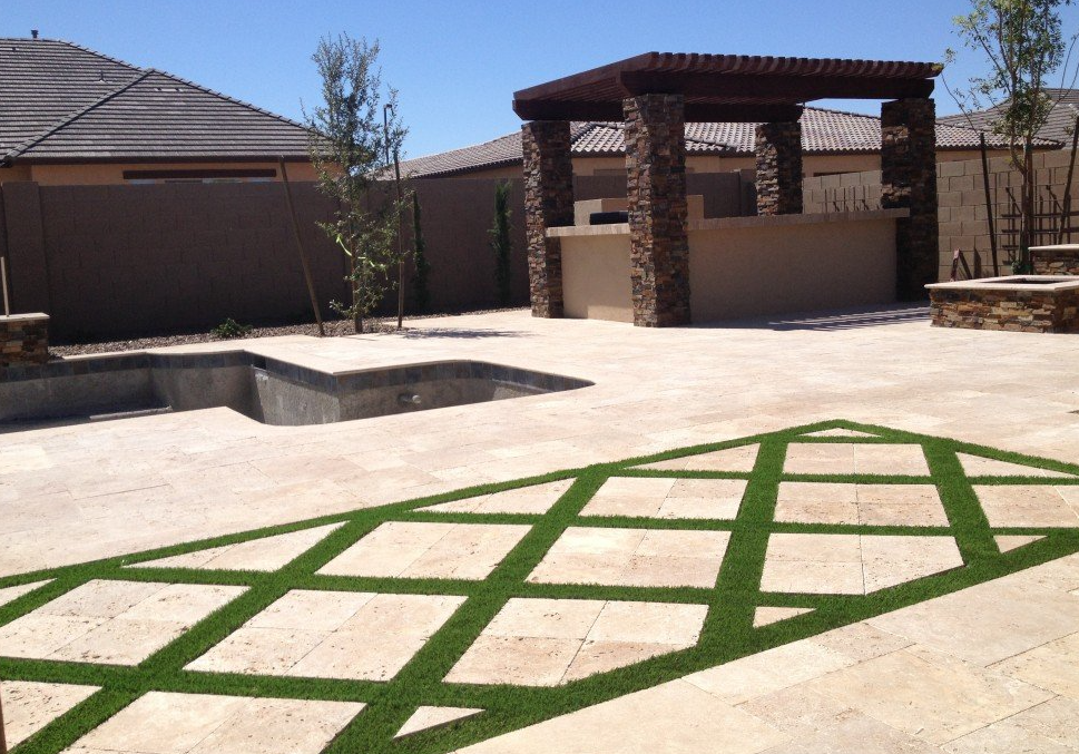 A backyard patio featuring a stone pavilion with wooden beams, a countertop, and a grill. The area showcases beige stone paving with a geometric grass pattern, including Scottsdale artificial grass for waterless lawns. An unfilled swimming pool is visible, surrounded by desert landscaping and block wall fencing.