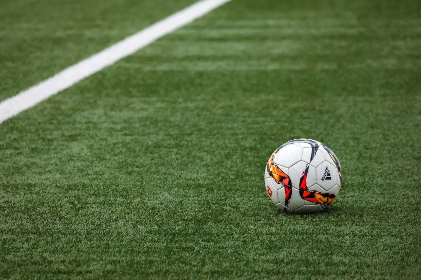 A soccer ball with a white base and a red, yellow, and black design rests on the green artificial turf of one of our premium playing fields near a white boundary line.
