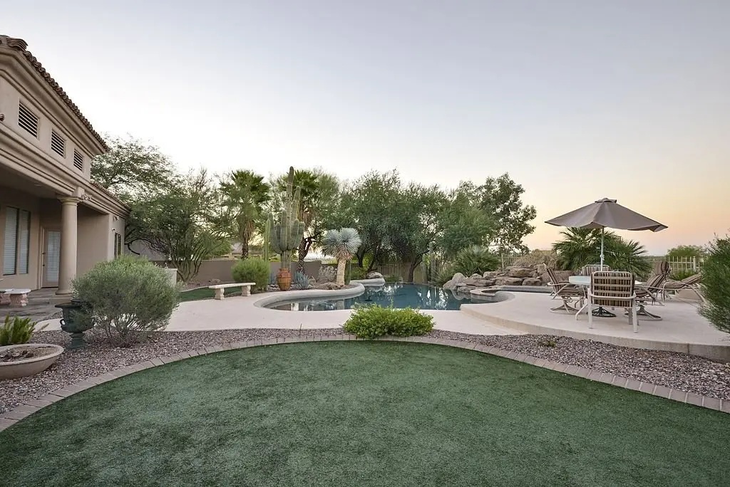 A luxurious backyard in Tempe, AZ features a curved swimming pool surrounded by lush greenery and palm trees. There's a patio area with a table, chairs, and an umbrella. Nearby, top-quality artificial grass installation complements the serene atmosphere, with a house visible on the left.