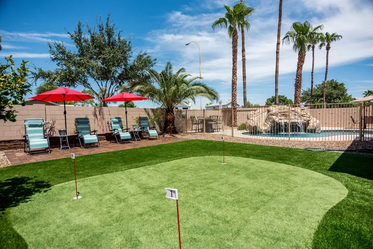 A sunny backyard in Chandler, AZ features a quality putting green in the foreground, a pool with a rock waterfall, lounge chairs with red umbrellas, and tall palm trees in the background. Blue skies and scattered clouds complete the relaxing scene. For installation, contact local artificial putting green installers.