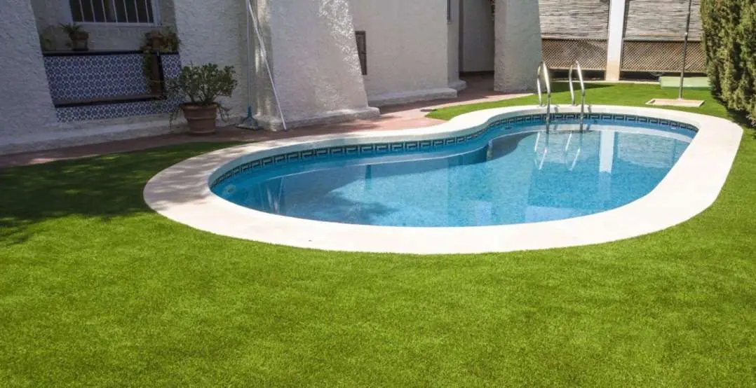 A small, oval-shaped swimming pool with clear blue water is surrounded by lush artificial grass lawns. The pool has metal handrails and is adjacent to a white-painted building adorned with plants in pots and outdoor tiling, all meticulously maintained by licensed and insured landscape pros from the East Valley Area.