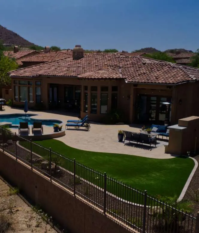 A luxurious single-story home in Gilbert, AZ, with a large patio and swimming pool. The house boasts a terracotta tile roof and multiple windows. The backyard features experienced artificial grass installers' work, lounge chairs, and a landscaped area, all enclosed by a decorative wrought-iron fence.