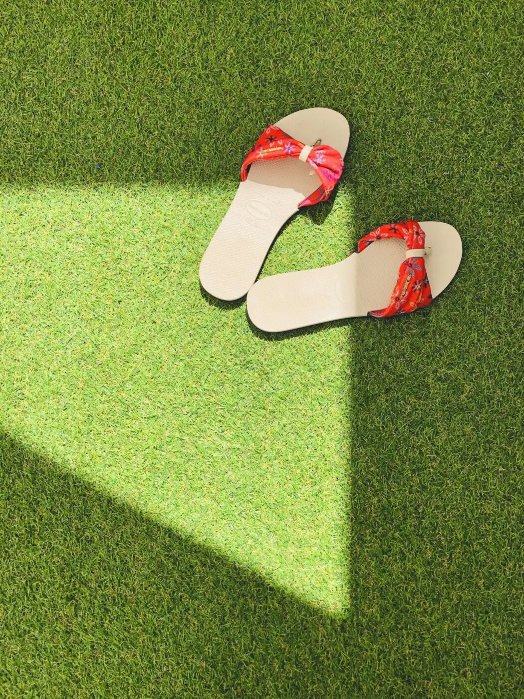 A pair of beige flip-flops with red patterned straps rests on an expanse of artificial grass, with a triangular shadow partially covering them.