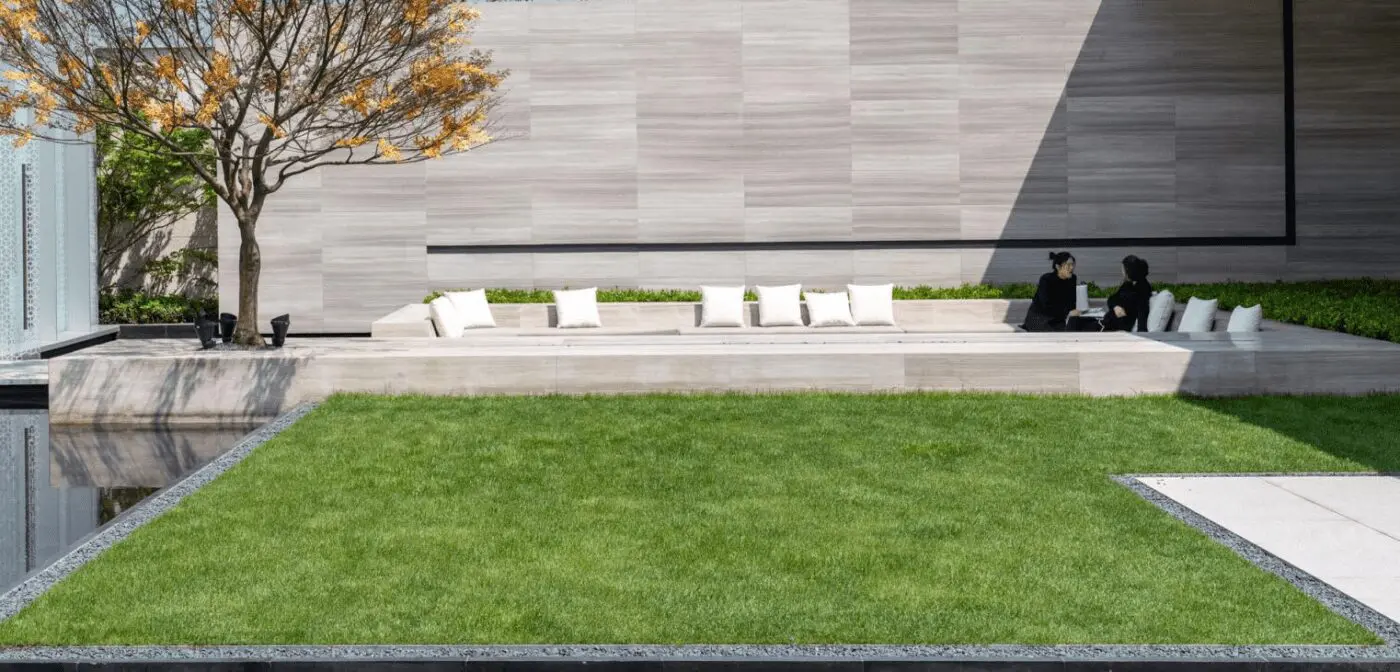 A modern outdoor space in Tempe AZ features a minimalistic design with an artificial grass installation, concrete pathway, and a large, bare tree to the left. At the far end, there is a long, low concrete bench with white cushions and two people sitting nearby in East Valley, engaged in conversation.