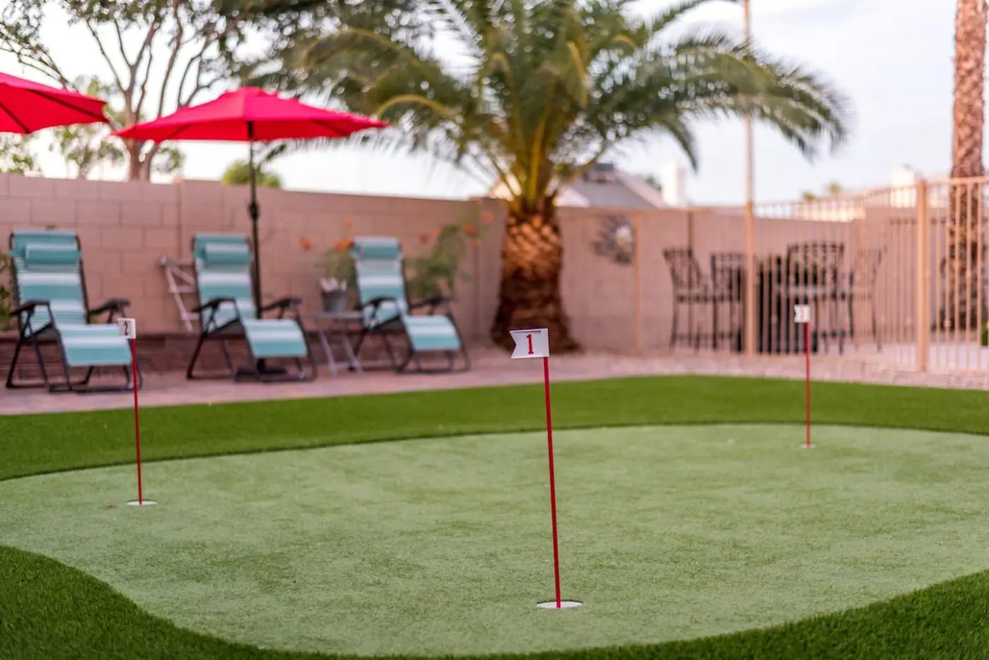 A backyard artificial putting green with three numbered flags. Lawn chairs with blue and white stripes and red umbrellas are set up on a patio area. A palm tree and a brick wall border the yard in Chandler, AZ. There is a metal table with chairs in the background, ensuring quality relaxation spaces.