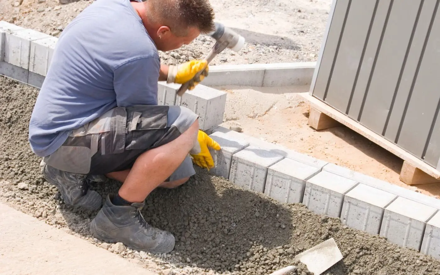 A construction worker, wearing a blue shirt, shorts, and yellow gloves, uses a hammer to align concrete blocks while kneeling on a gravel surface. The scene showcases East Valley xeriscape techniques amidst the sand, gravel, and partially built structure perfect for desert landscaping.