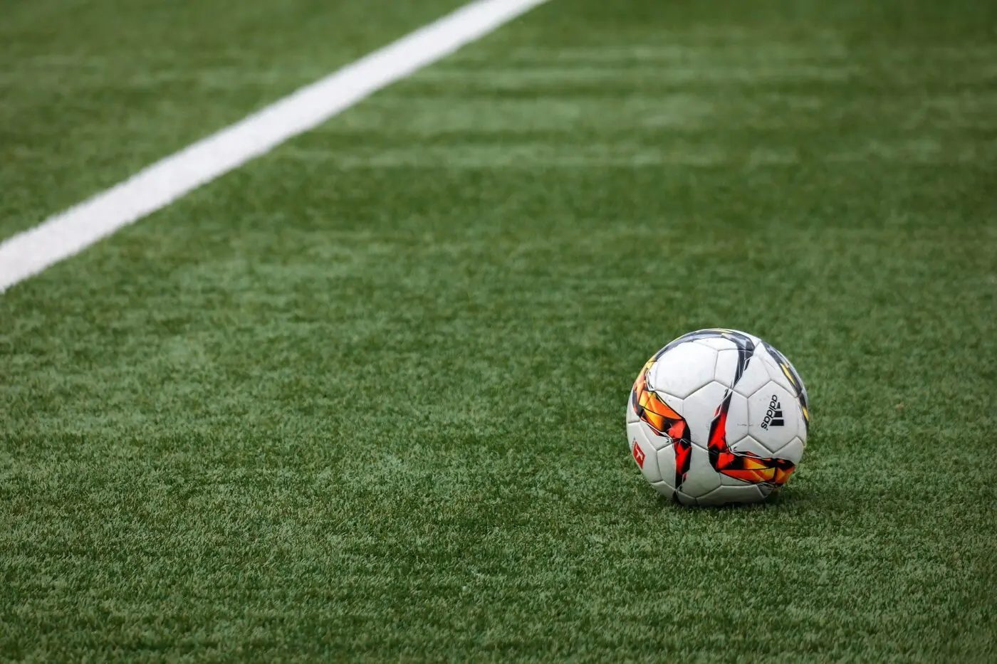 A soccer ball with colorful patterns rests on the green artificial grass, near a white boundary line. The field in Queen Creek features neatly trimmed, bright green turf.