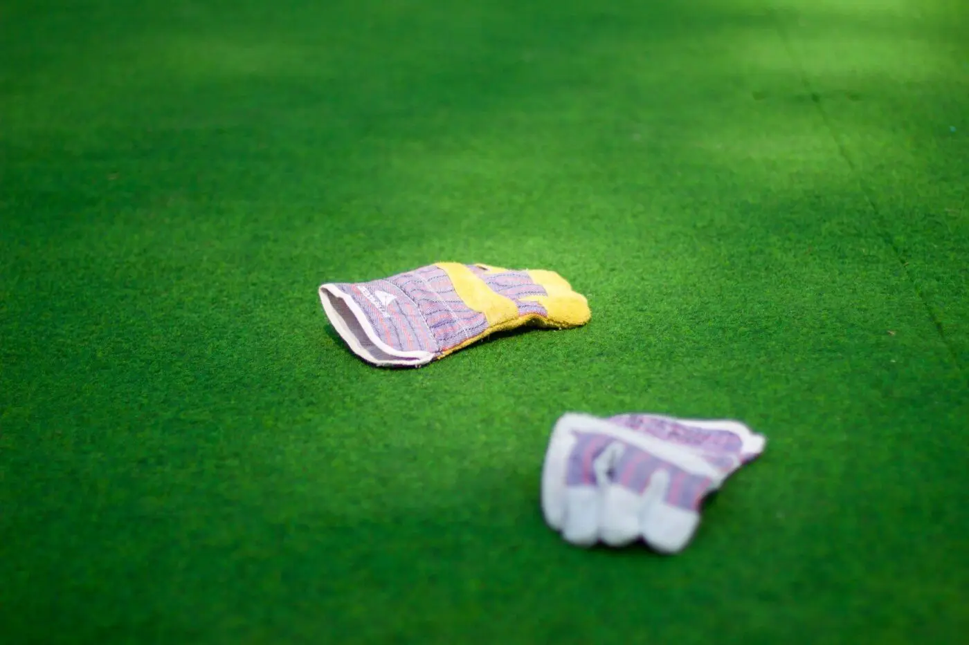 Two work gloves lie on vibrant artificial grass for playgrounds. One glove is positioned closer to the camera and appears partially open, while the other is further back, laying flat. The gloves are multicolored with shades of purple, yellow, and white.