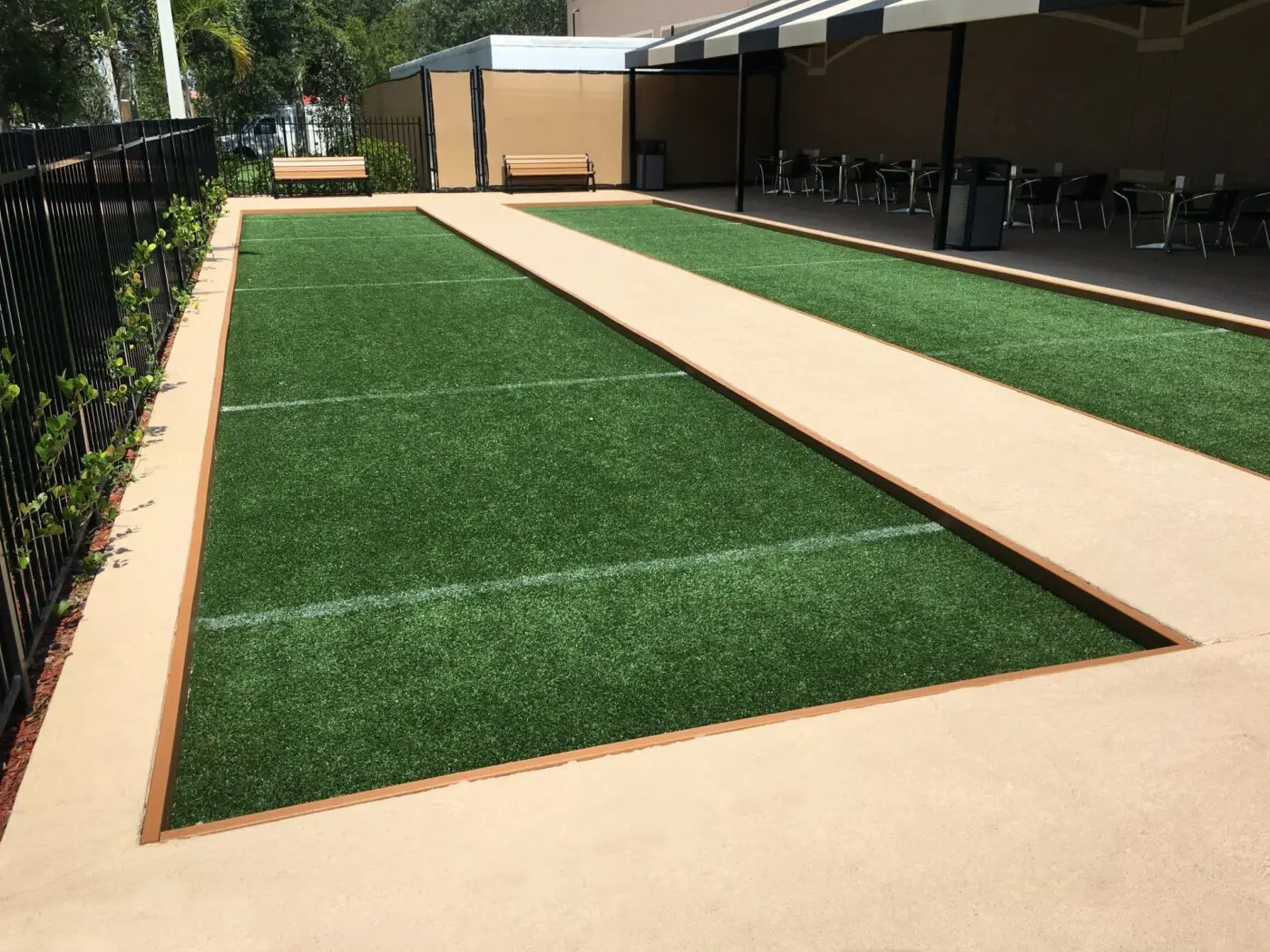 A clean, well-maintained bocce ball court with artificial turf is surrounded by low beige barriers. Adjacent to a patio area with tables and chairs under shade coverings, it offers a relaxing spot to enjoy the game. A bench at the far end near a fence with climbing plants completes this inviting playing field.