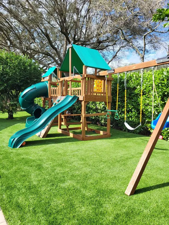 A small, simple model of a wooden house with a gabled roof sits on vibrant artificial grass, reminiscent of the installations in Tempe AZ. The house is made from square wooden blocks, giving it a minimalist and handcrafted appearance. The lush grass is evenly spread in the background.