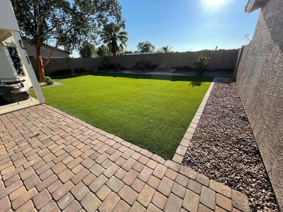 A backyard in Tempe AZ with a neatly trimmed rectangular lawn surrounded by stone pavers. To the left, there is a patio area with a barbecue grill, perfect for East Valley gatherings. A tree provides shade on the left side, and the yard, featuring an artificial grass installation, is bordered by a wooden fence and rocky landscape. The sky is clear and sunny.