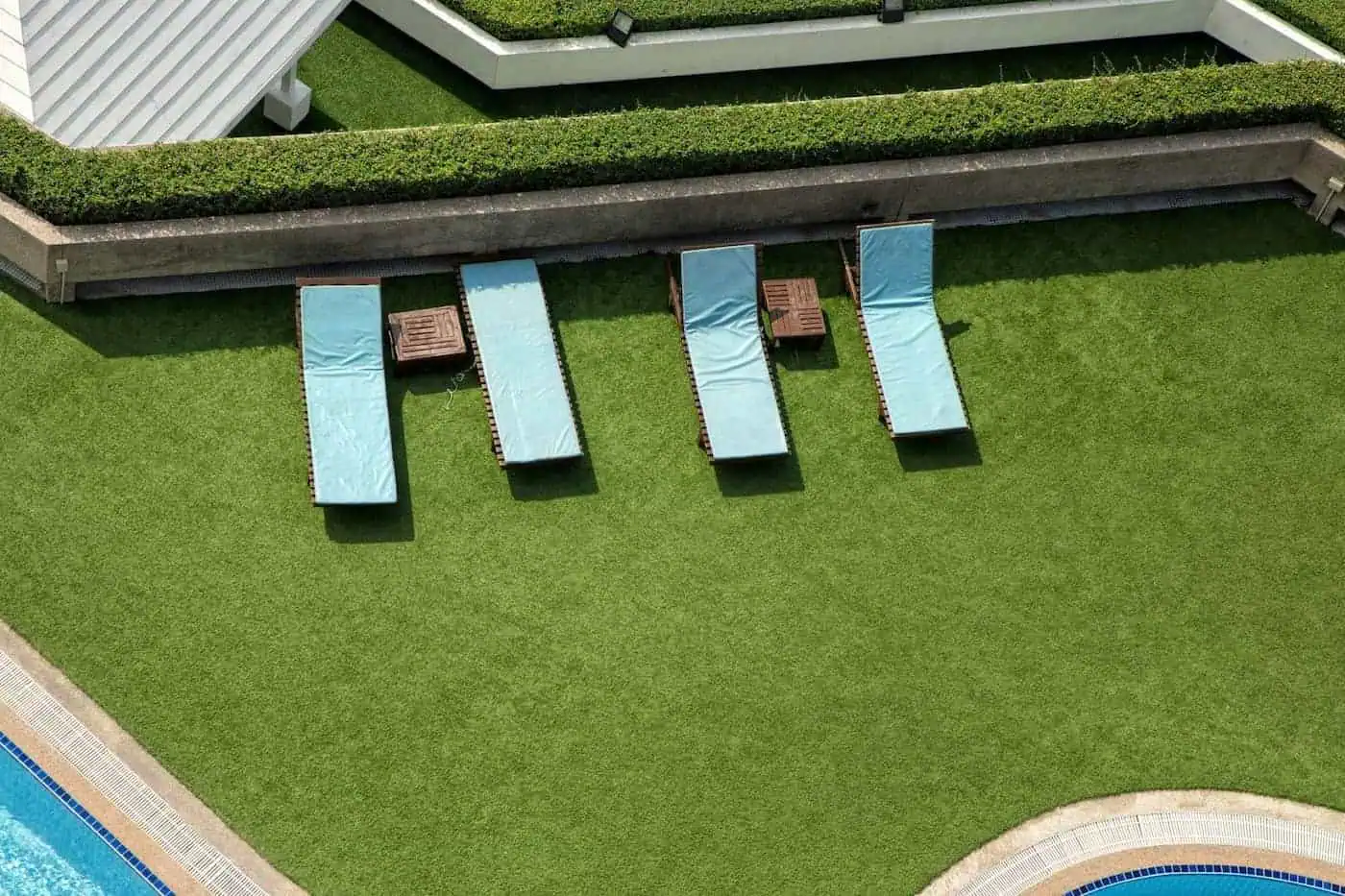 Four blue lounge chairs and three wooden tables are arranged on a manicured grassy lawn, next to a white building with green hedges. The scene, featuring pet-friendly turf installed by licensed & insured artificial grass professionals, appears to be part of a relaxing outdoor area near a pool with blue tiles visible in the bottom right corner.